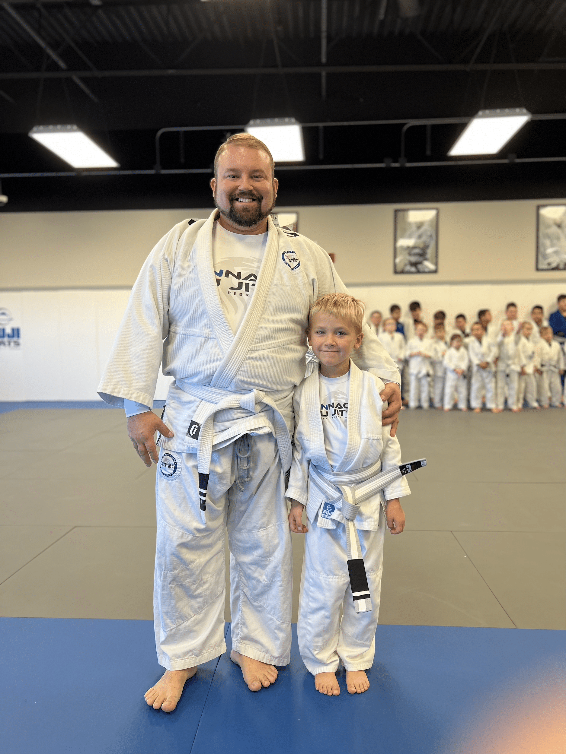 pinnacle jiu jitsu kid and adult in white gi posing for photo with class behind them